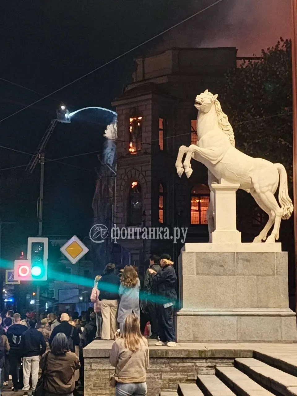 Пожарные проливают горящее здание с высоты пожарной вышки; пламя на нижних этажах постепенно становится слабее.